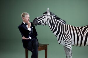 Max Raabe sitzt auf einem Tisch neben einem Zebra